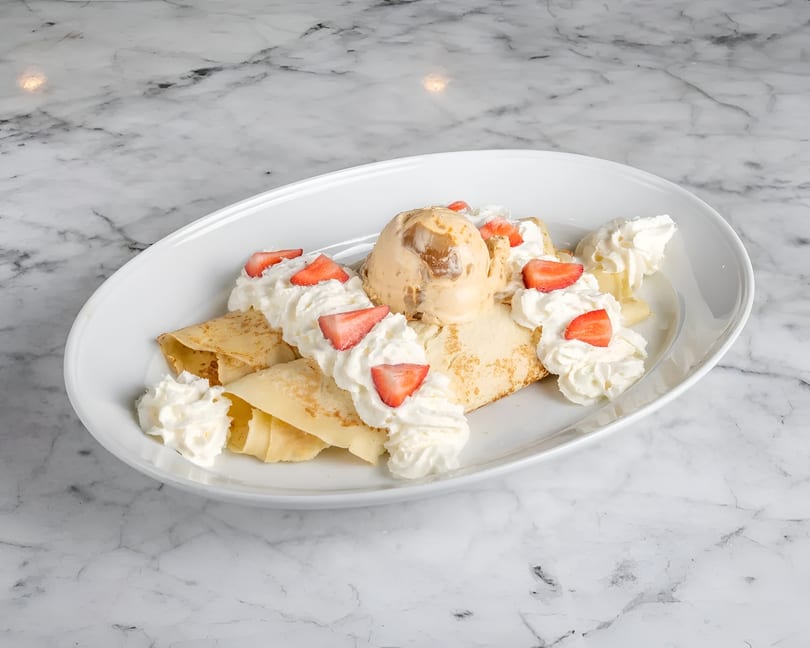 Crepes topped with a scoop of caramel ice cream, whipped cream, and sliced strawberries on a white plate.